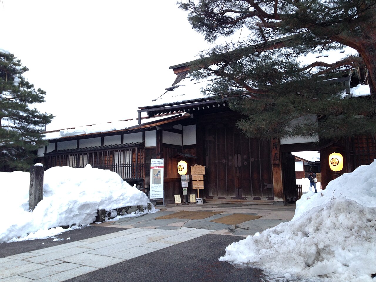 The wooden main gate and guardhouse row of Takayama Jinya under snow, with pines behind and snowbanks piled along the paved approach.