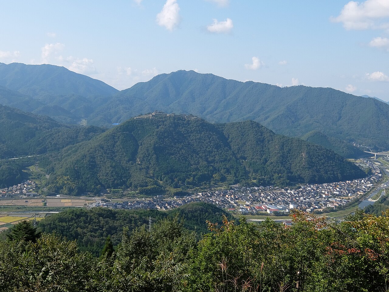 View across a valley town to a forested hill with the Takeda Castle ruins on its summit, under a blue sky with scattered clouds.