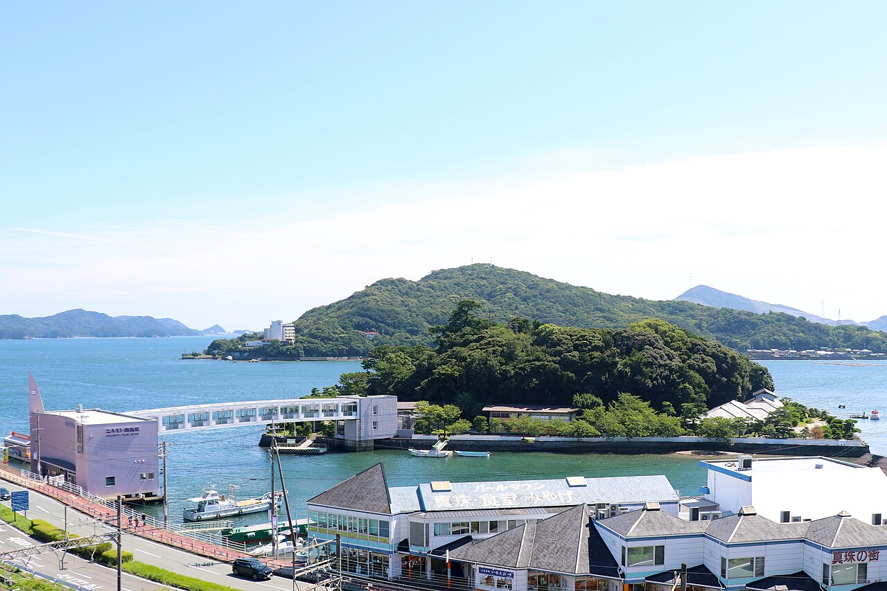 A small wooded island with a museum building on its shore, seen across the blue water of Toba Bay under a clear sky