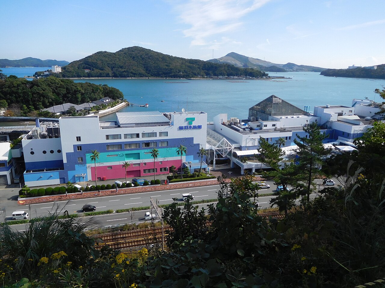 A hillside view over a green island, a bay and the waterfront buildings of Toba port under a blue sky