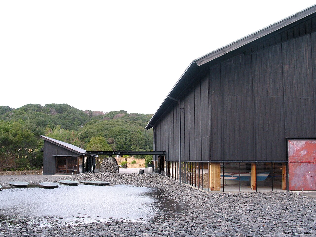 The dark timber exterior of the Toba Sea-Folk Museum with a stone-paved forecourt and wooded hills behind