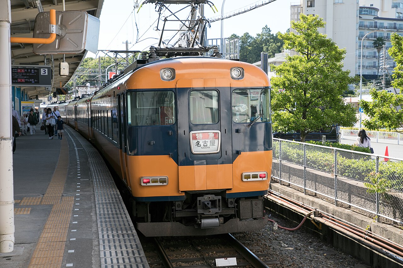 An orange Kintetsu limited express train stopped at a platform at Toba Station beside a tree and apartment blocks