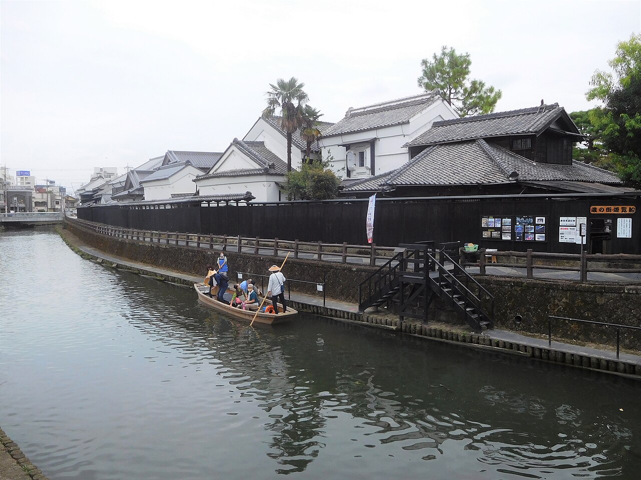 A small wooden sightseeing boat with a boatman in a sedge hat on the Uzuma River in Tochigi, below a long black wooden fence and white-walled storehouses