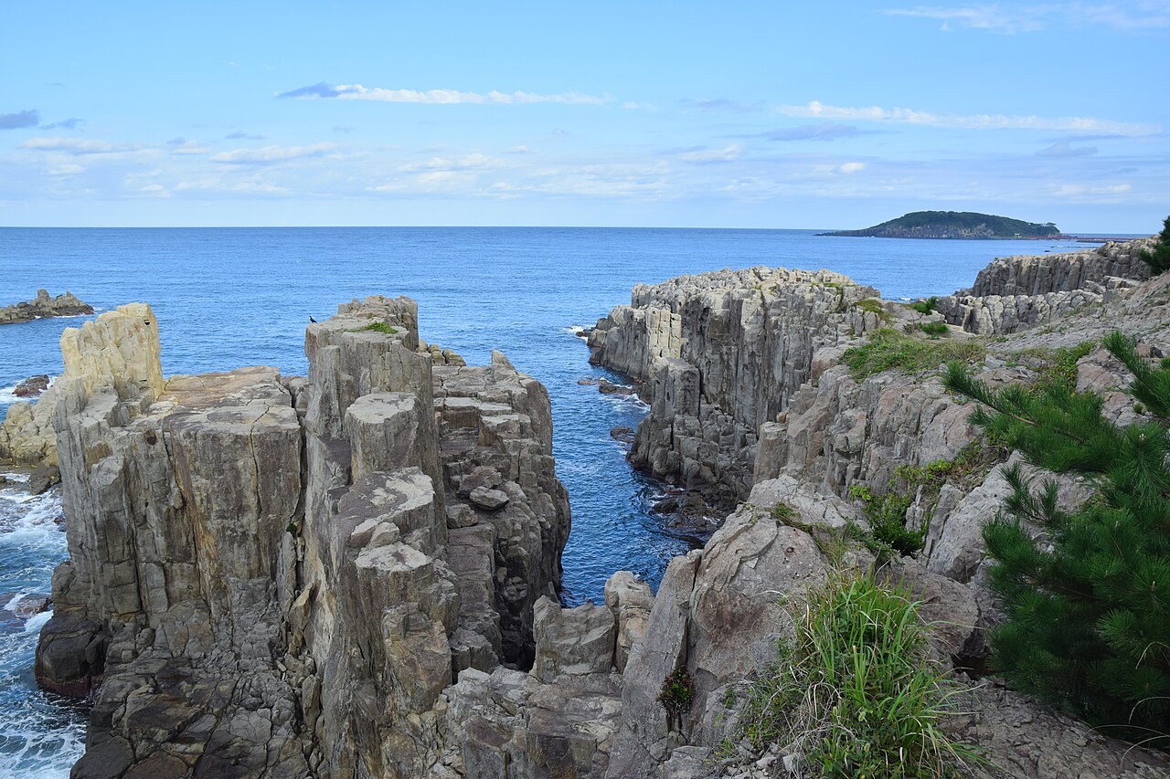 Tall columns of grey rock at the Tojinbo cliffs above a calm blue sea, with Oshima island low on the horizon