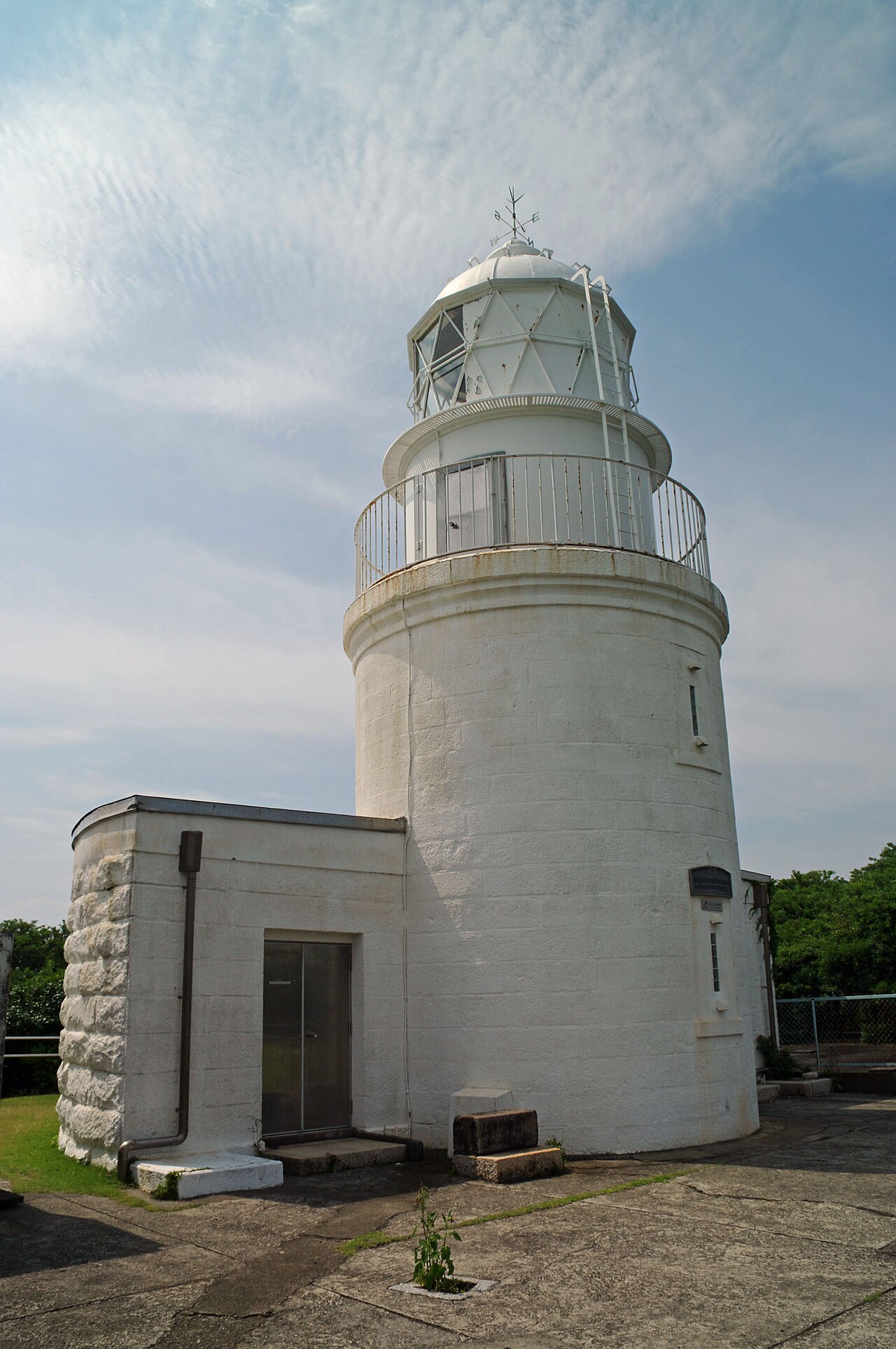 The white Tomogashima Lighthouse tower with its small attached building on a hilltop under a partly cloudy sky.