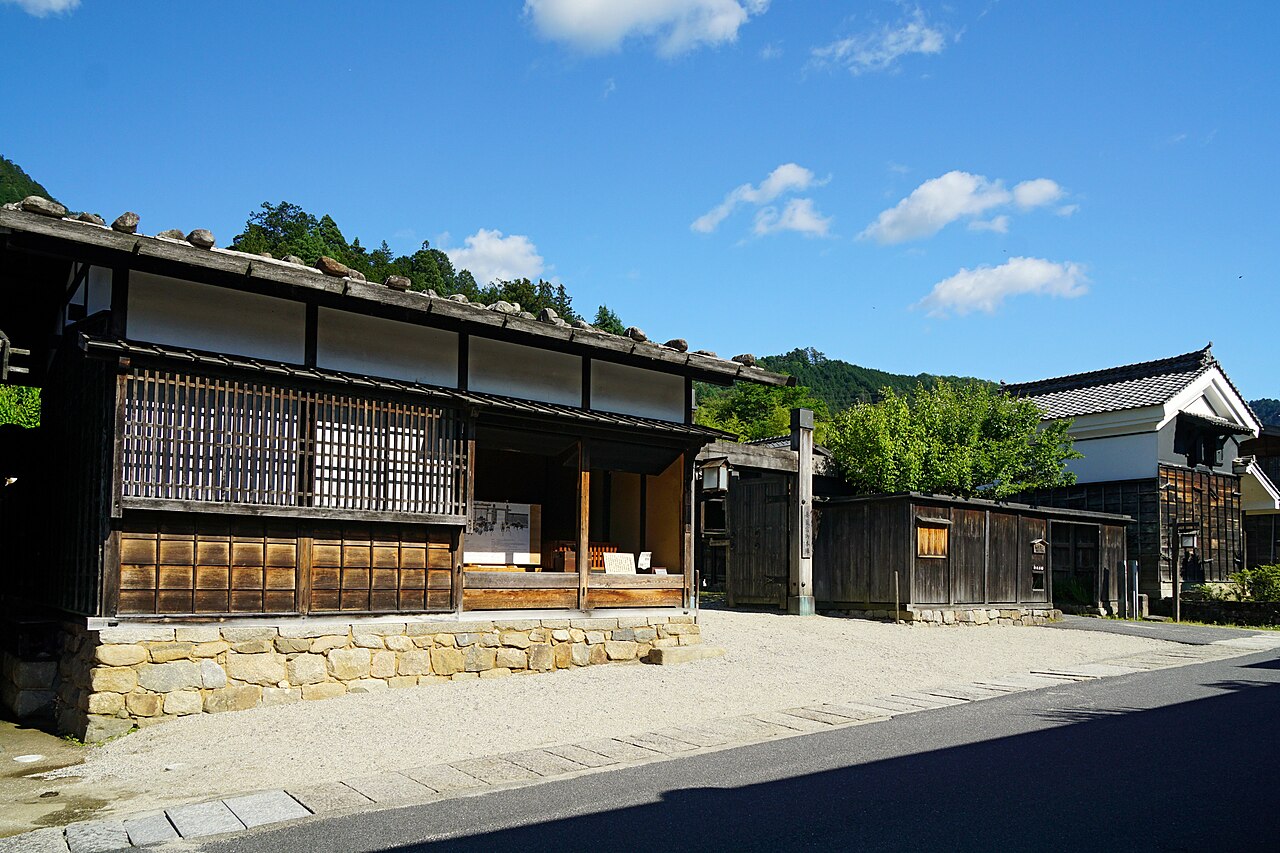 A low wooden building with a stone-weighted roof beside the street, a gravel yard in front and mountains behind.