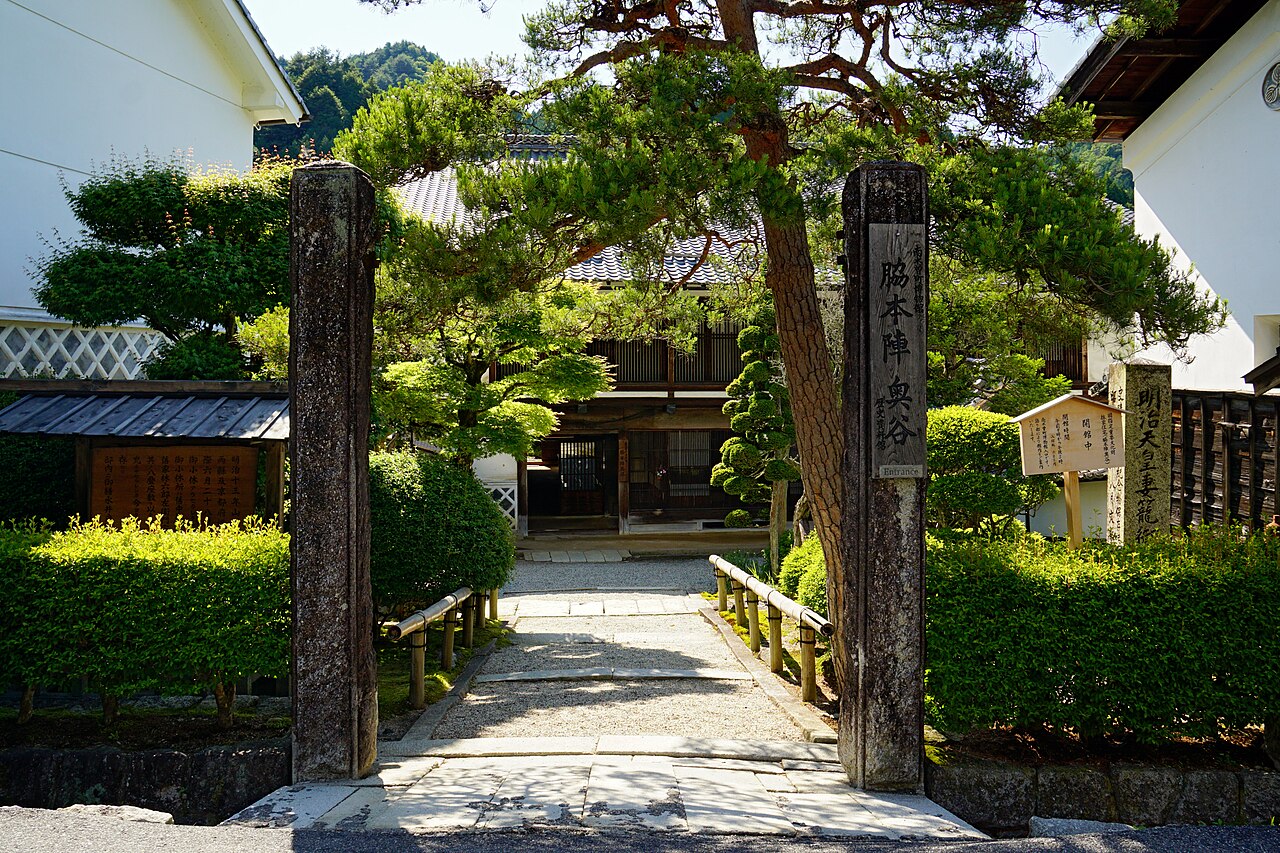 Stone gateposts inscribed Waki-honjin Okuya, a gravel path with pine trees, and a wooden house beyond.