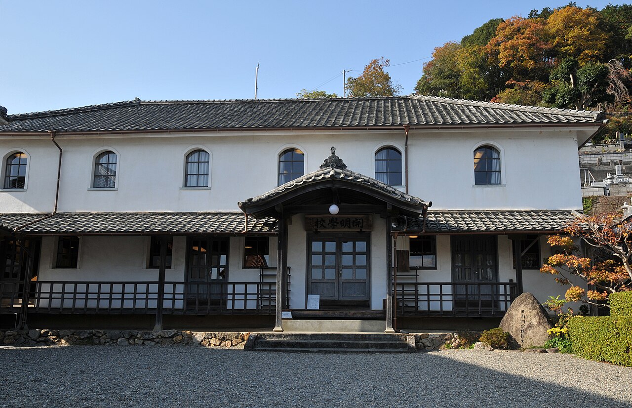 The white-plastered, tile-roofed wooden Kaimei School building with a gabled entrance porch and a gravel yard, set against autumn hills in Uwa, Seiyo.