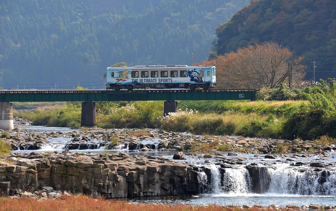 A white Wakasa Railway railcar crossing a bridge above a rocky river with a low waterfall, autumn colors on the hillside