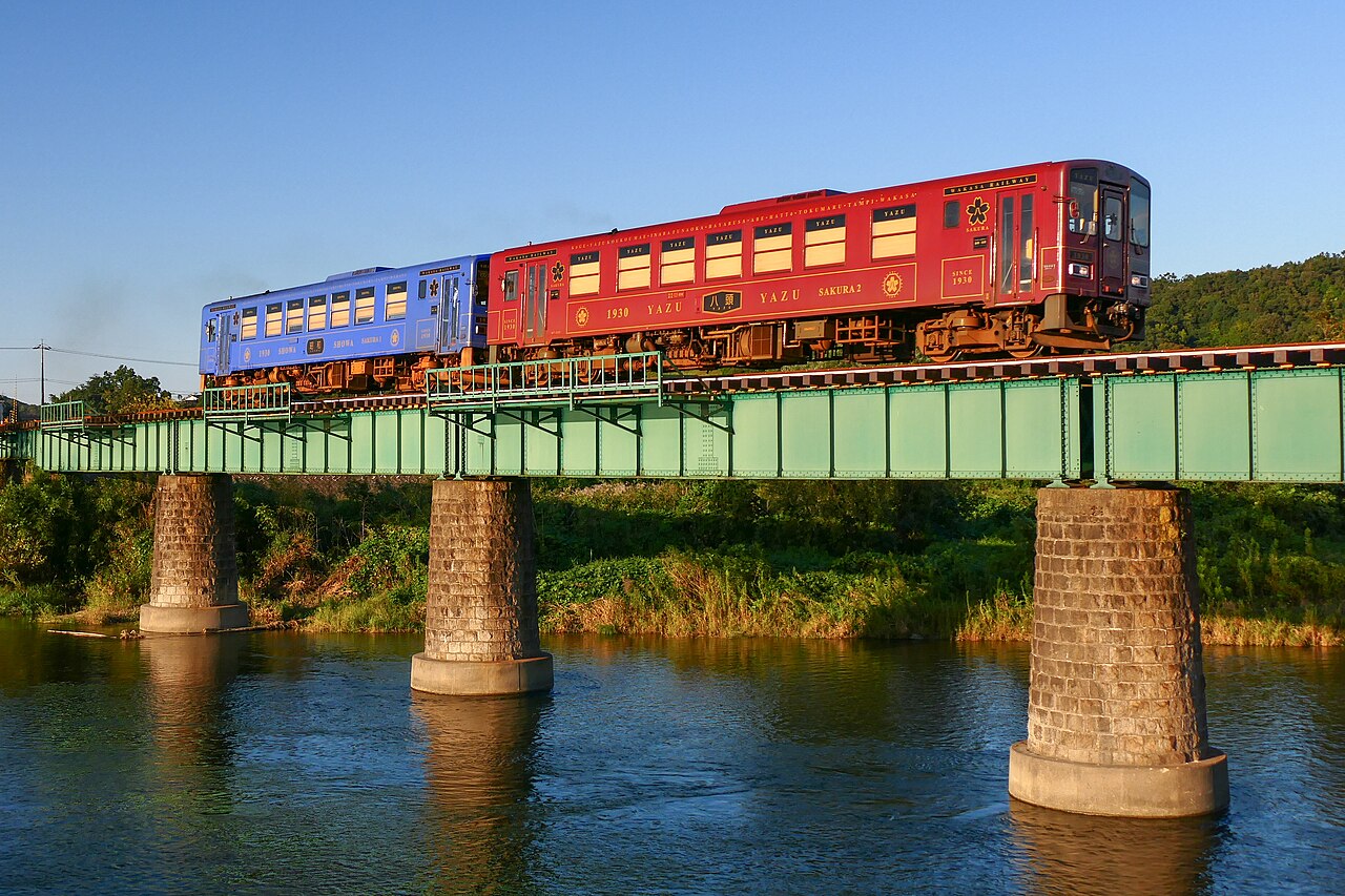 A red and a blue Wakasa Railway railcar crossing a green girder bridge over a river in evening light