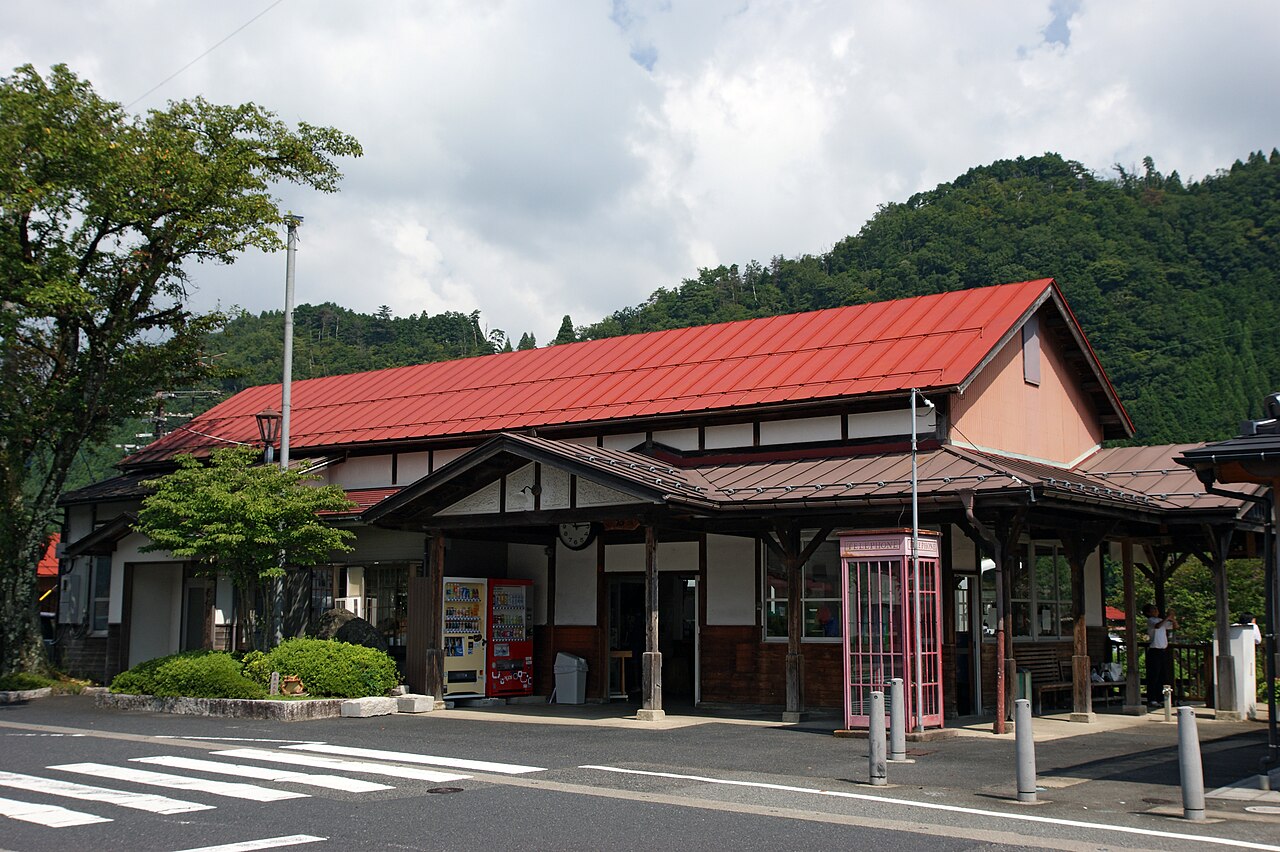 The wooden Wakasa Station building with a red roof, a pink telephone box and wooded hills behind