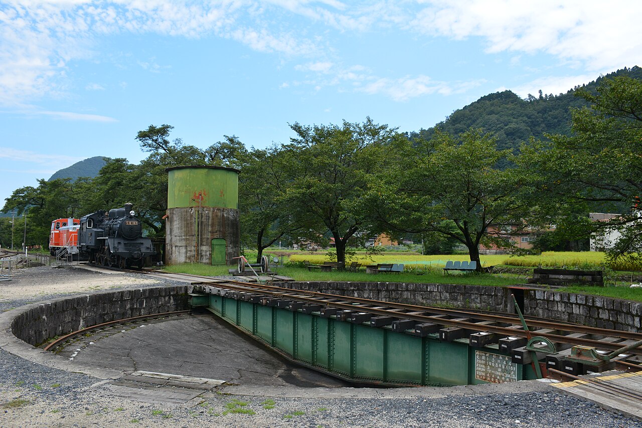 A black steam locomotive and an orange diesel locomotive stand beside a green water tower, with the turntable in the foreground at Wakasa Station