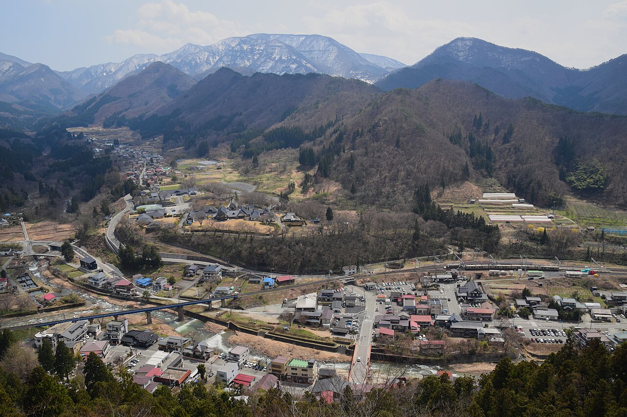 The small town of Yamadera and its river seen from the Godaido viewpoint high above, with snow-patched mountains beyond
