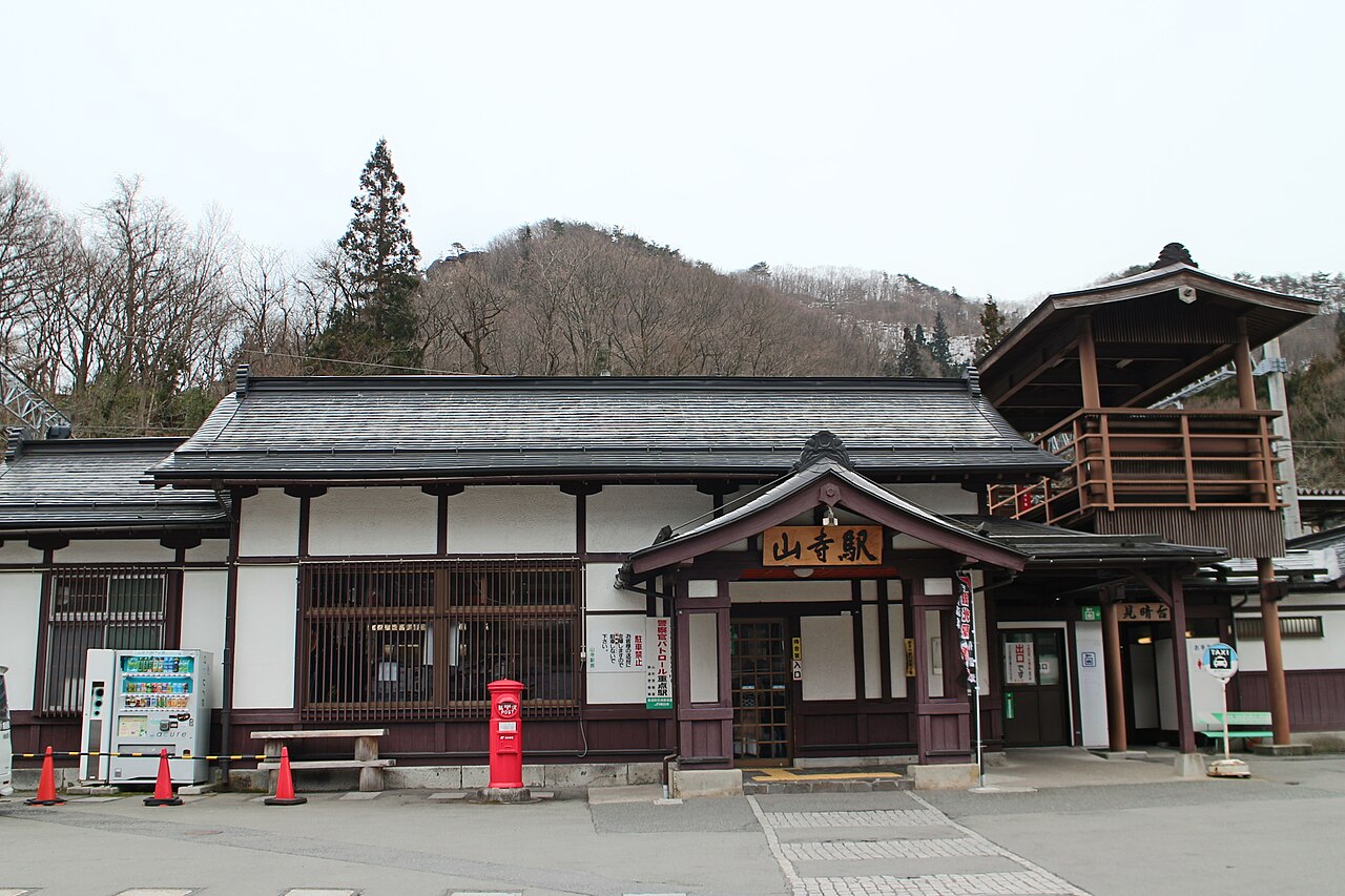 The traditional wooden station building of JR Yamadera Station with its lookout tower and red post box, a hill rising behind