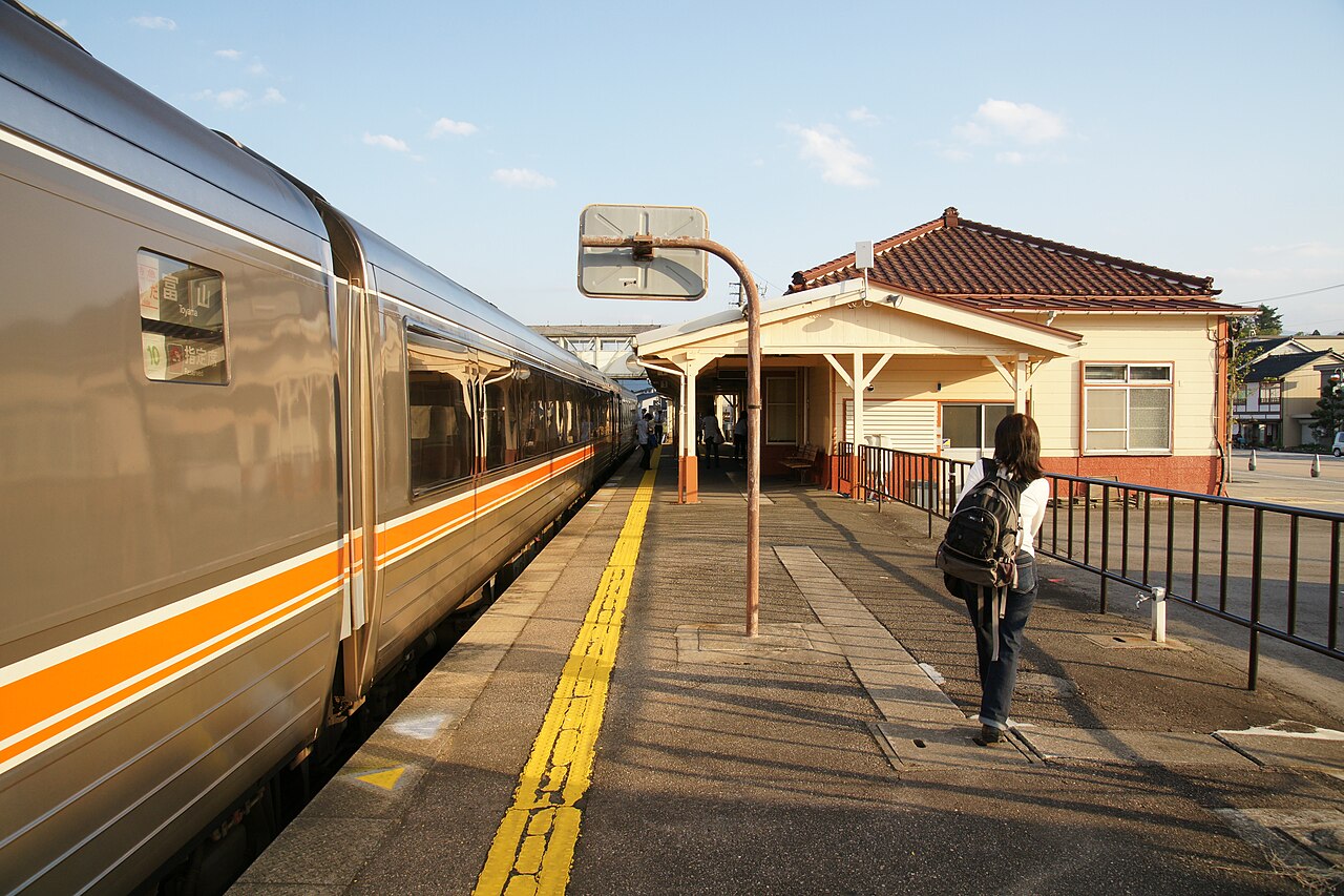 Platform at Etchu-Yatsuo Station with a stopped train on the left, a traveler with a backpack walking ahead, and the low tile-roofed station building on the right.