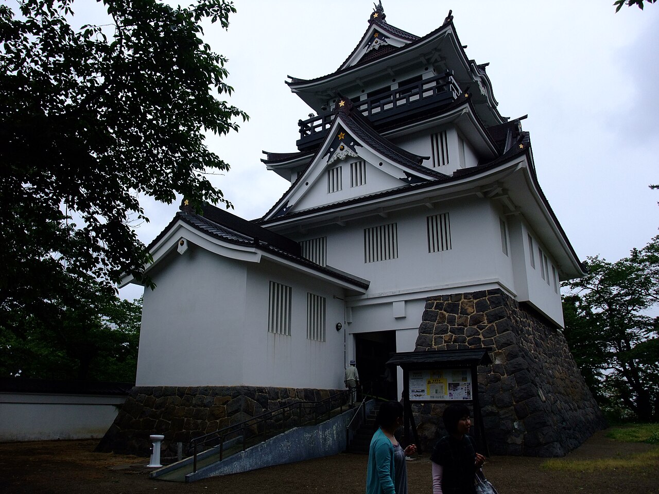 A white-walled castle keep with layered tiled roofs on a stone base, framed by trees.