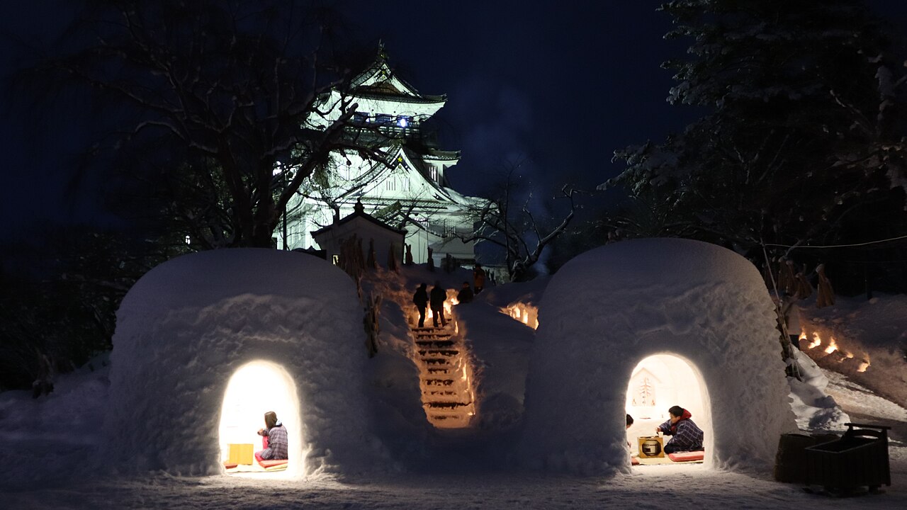 Snow-covered kamakura huts glowing at night with a lit castle keep rising behind them.