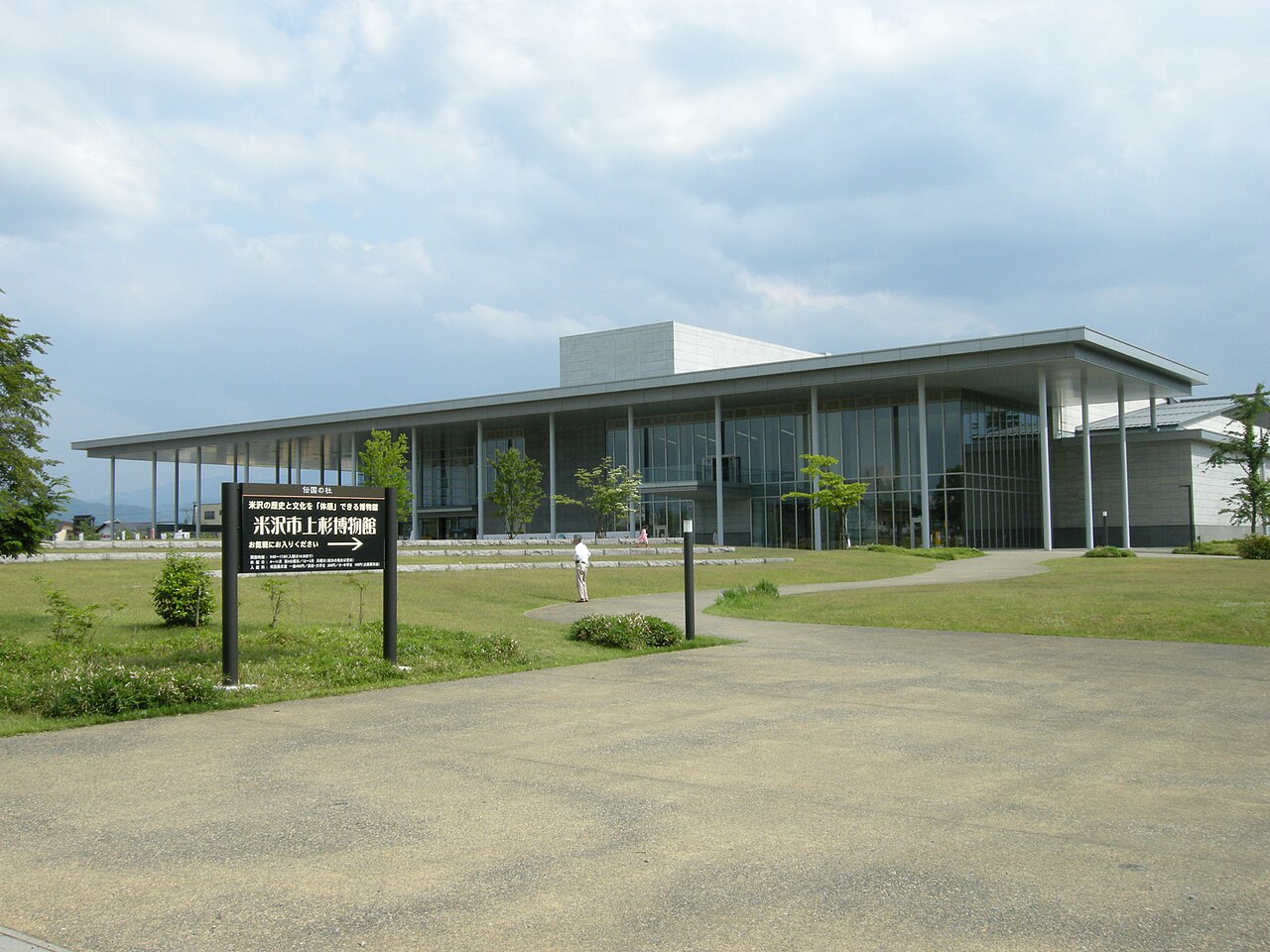 The long, low-roofed glass-fronted building of the Yonezawa Uesugi Museum beyond a lawn and a signboard, under a cloudy sky.