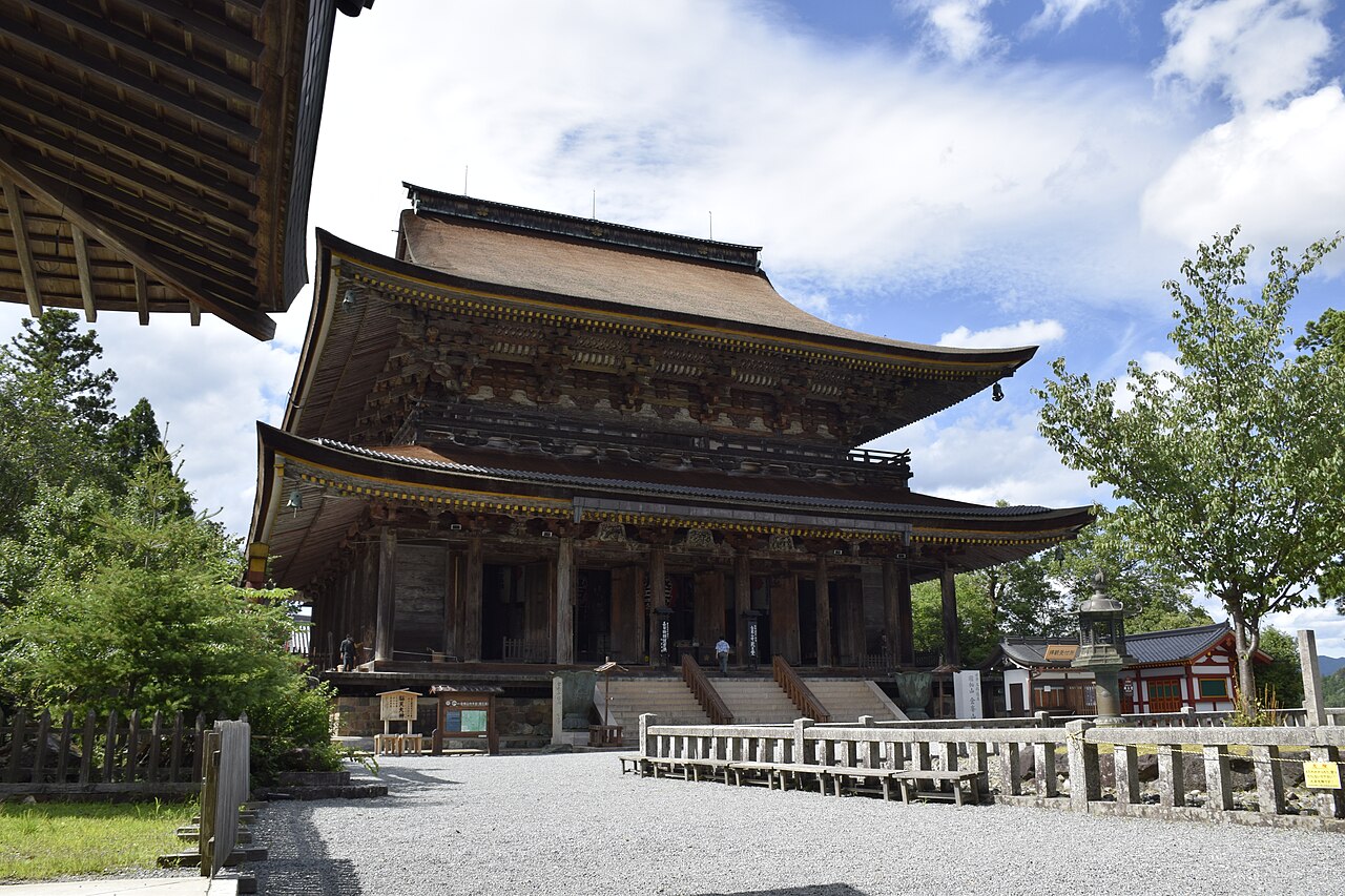 The massive dark-timbered Zao-do main hall of Kinpusen-ji temple in Yoshino, seen across a gravel courtyard under a partly cloudy sky, with green trees beside it