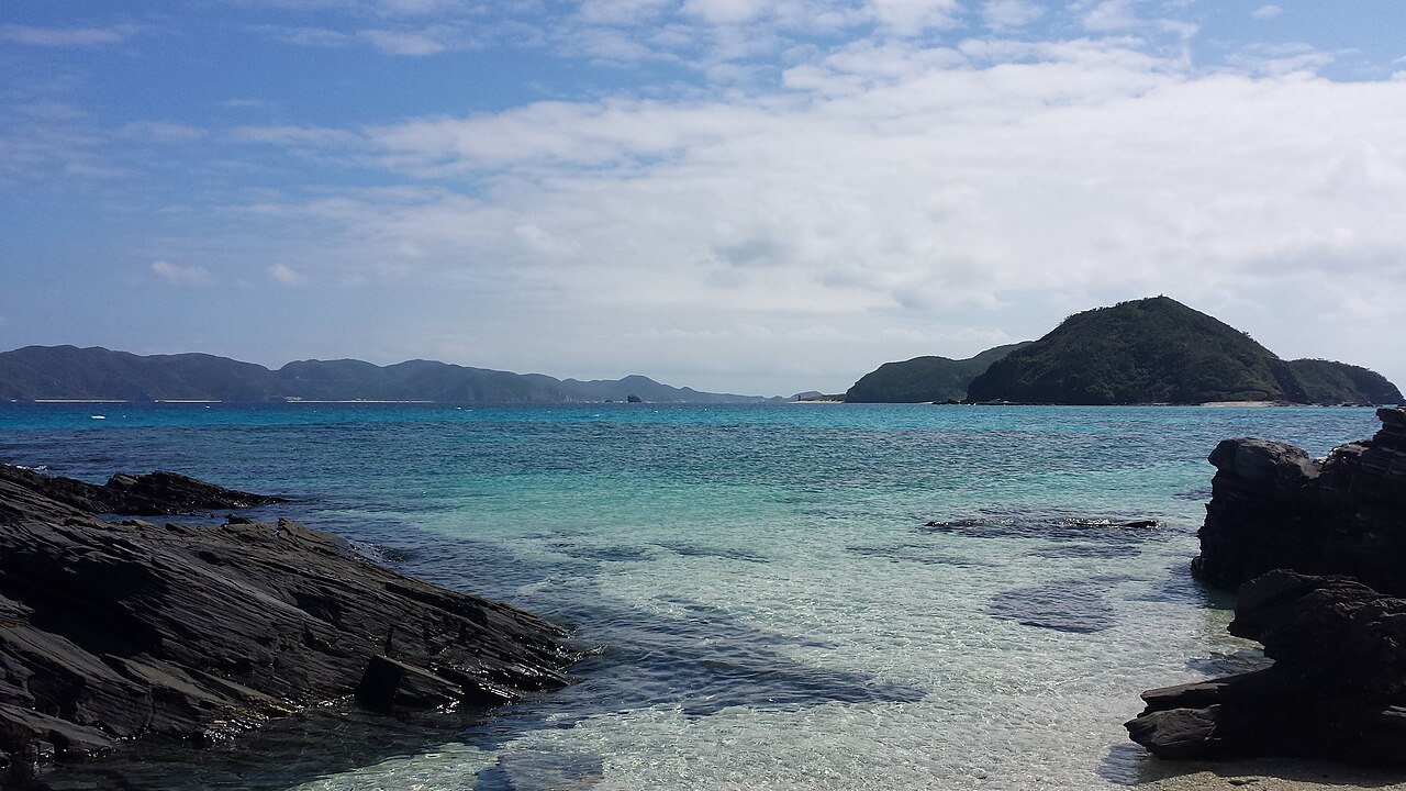 Clear shallow water over a rocky shore on Zamami Island, with distant Kerama islands under a pale sky.