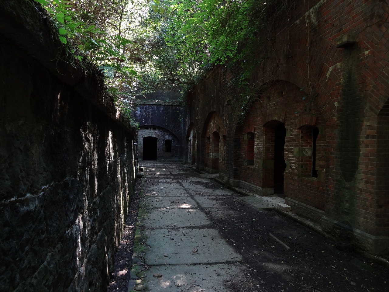 A paved passage between a stone wall and arched brick chambers at a ruined battery, with leaves overhanging the path.