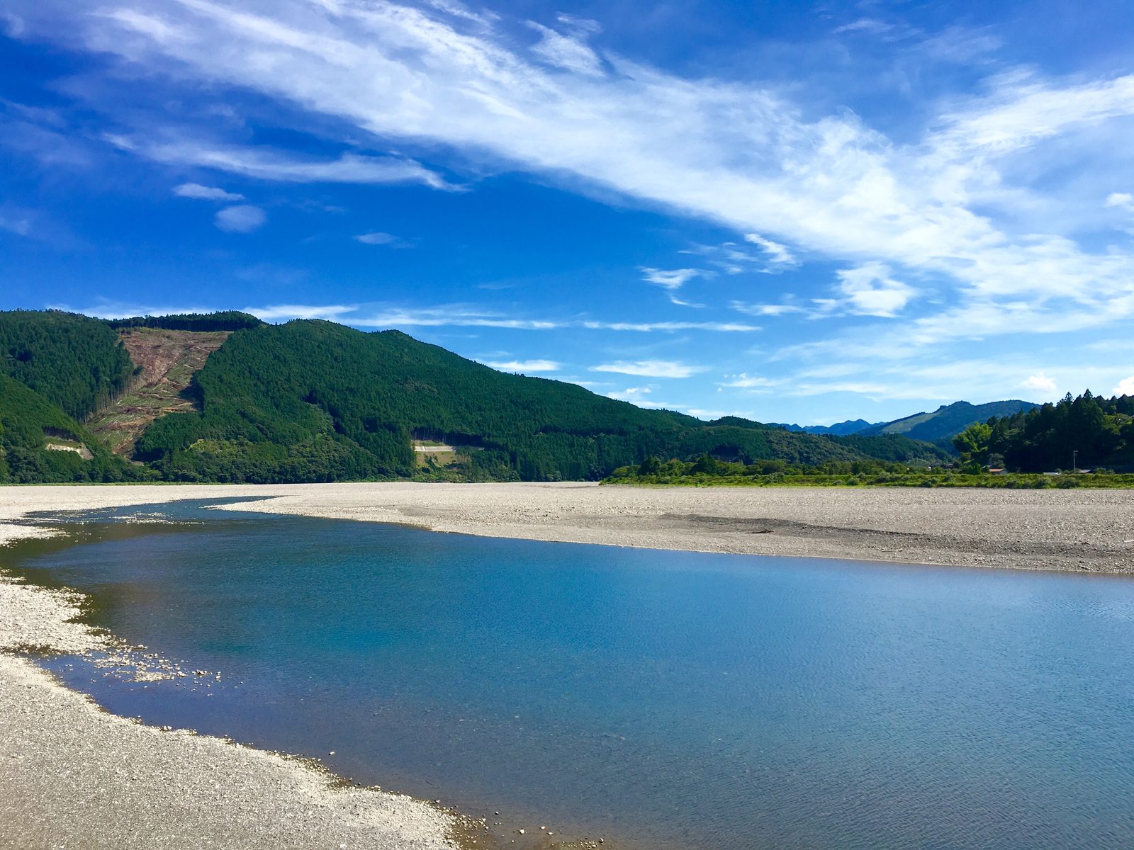 Wide gravel bar and a calm blue stretch of the Kumano River with forested green hills under a bright sky