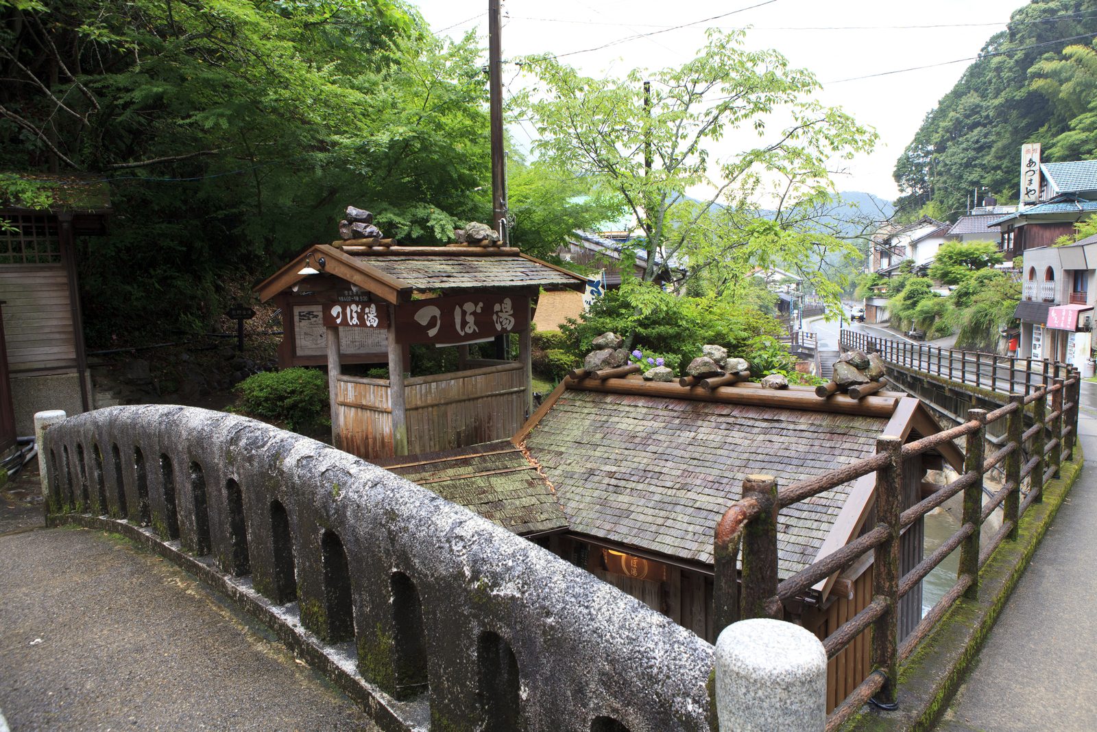 Shingle-roofed huts beside a stone bridge in Yunomine Onsen, with the village street running along the river in the valley