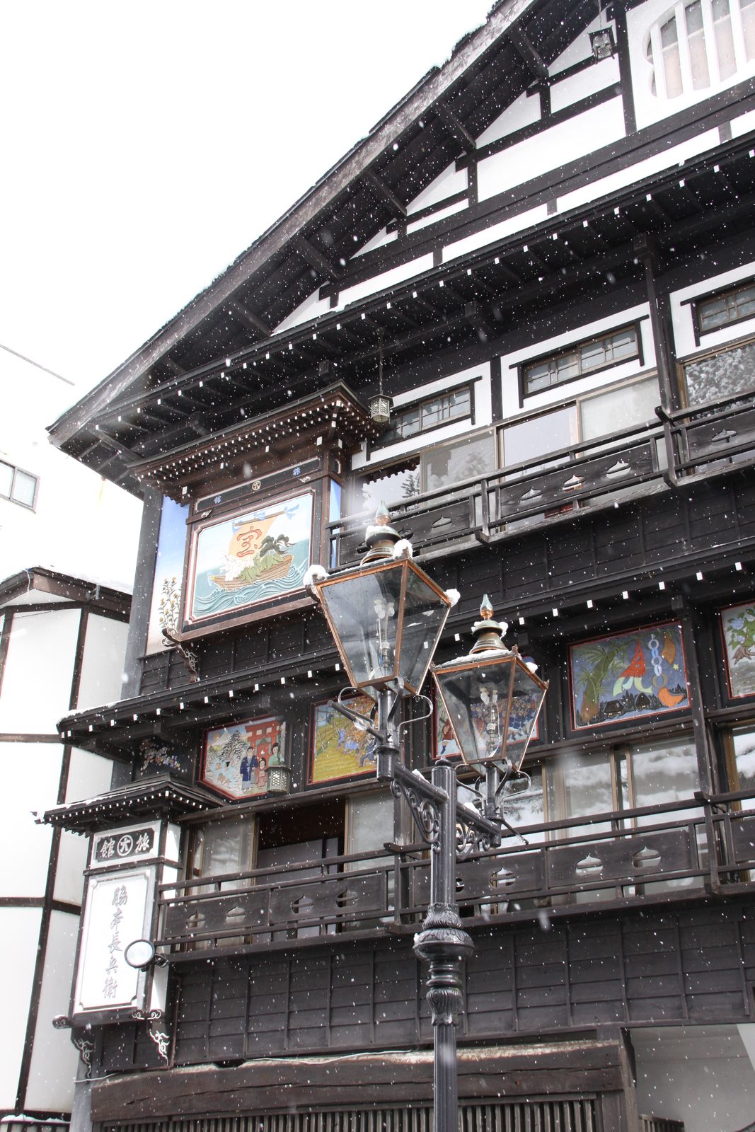 The front of a traditional multi-storey wooden inn with painted panels under the eaves and a gas lamp beside it, on an overcast snowy day.