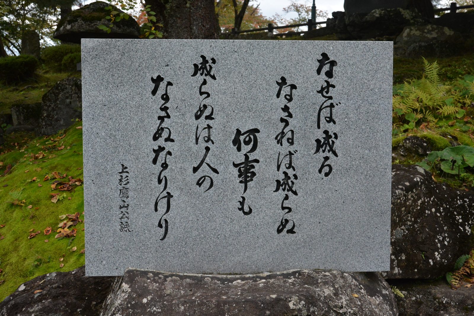 A grey granite monument carved with Uesugi Yozan's verse beginning "Naseba naru" in black calligraphy, set on mossy rocks at Uesugi Shrine in Yonezawa.