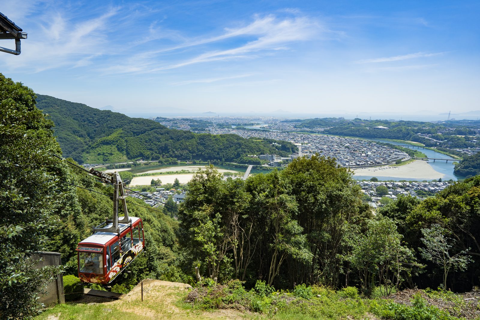 A red ropeway car above the forest with the Iwakuni townscape and the Nishiki River below
