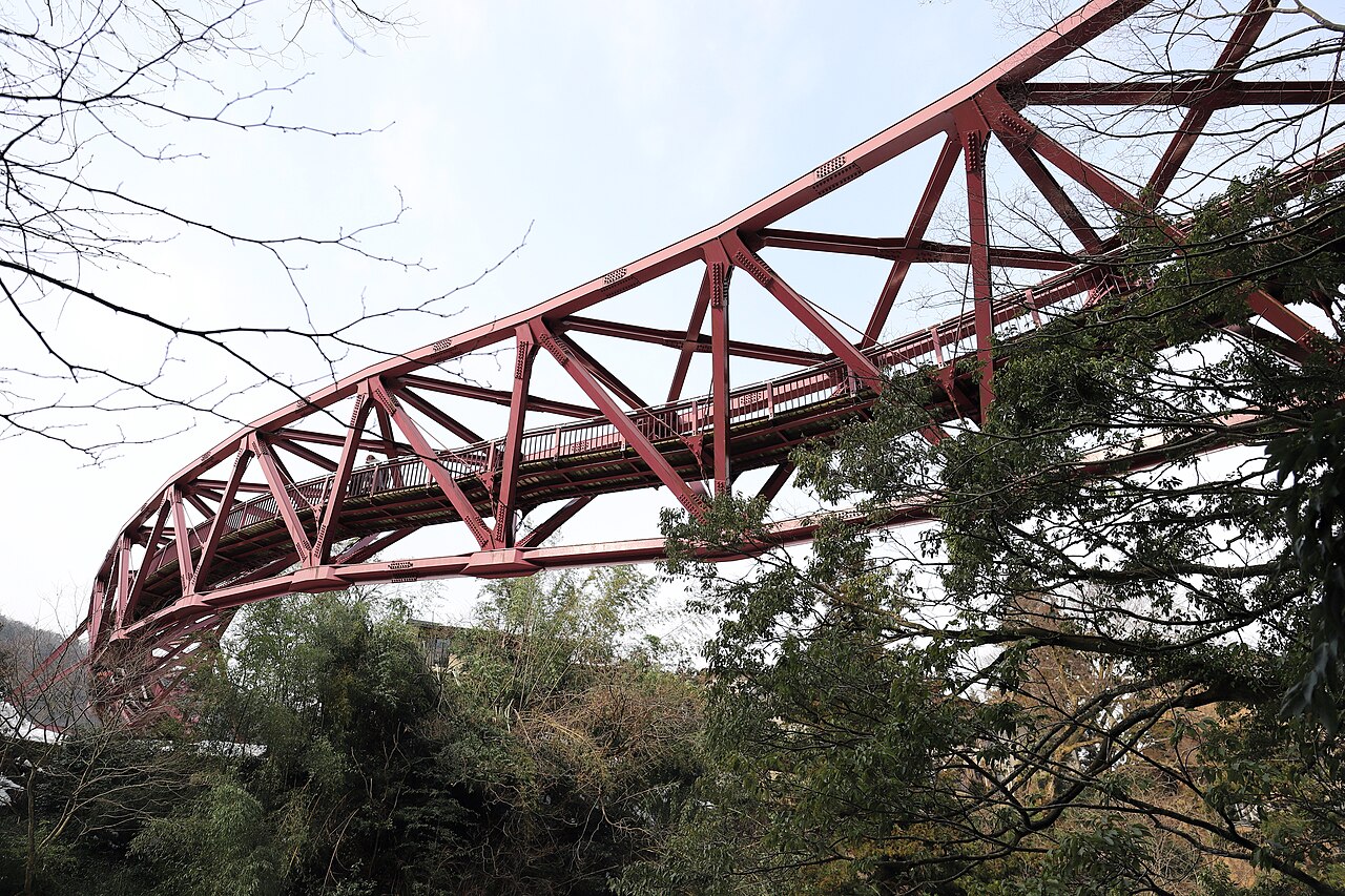The red steel truss of Ayatorihashi pedestrian bridge arching high above the wooded Kakusenkei gorge in Yamanaka Onsen, seen from below through bare tree branches.