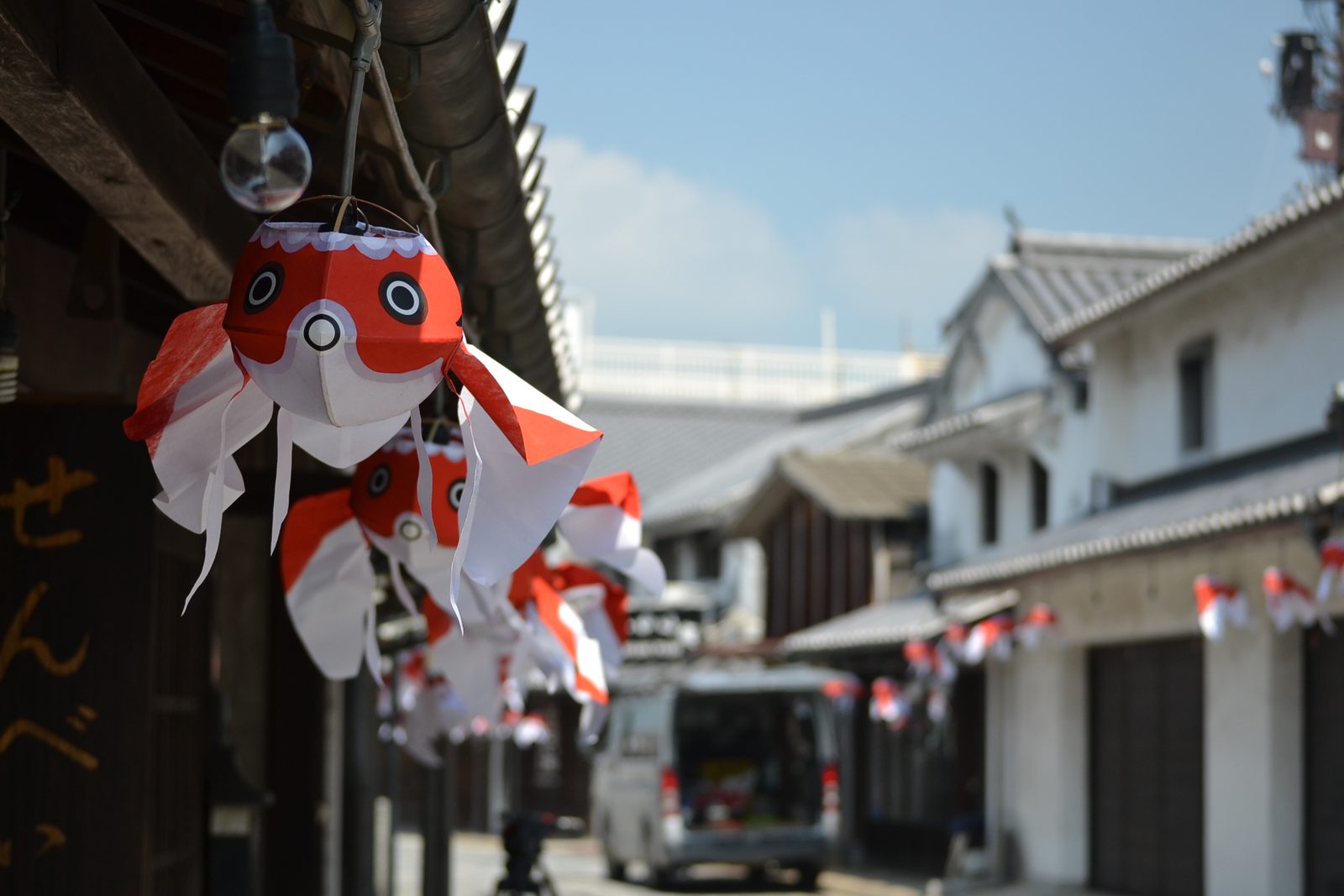 Close-up of a red and white paper goldfish lantern hanging from a wooden eave, with white-walled buildings blurred in the background